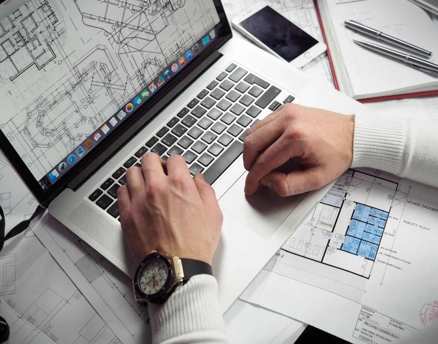 Hands typing on a laptop surrounded by printed plans and drawings